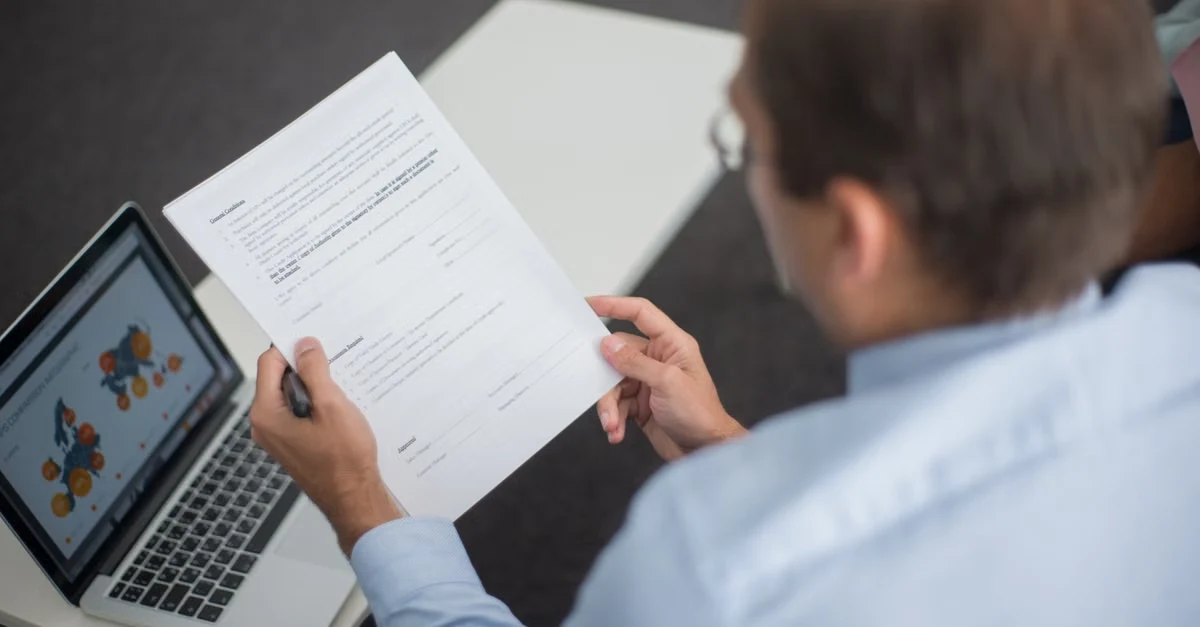 Executive reviewing printed document with laptop showing data analytics charts nearby on desk.
