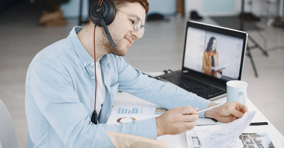Man wearing headphones reviewing documents at desk with laptop during video call.