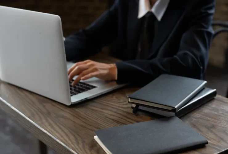 Professional in business suit typing on laptop at wooden desk with notebooks and contact database management materials.