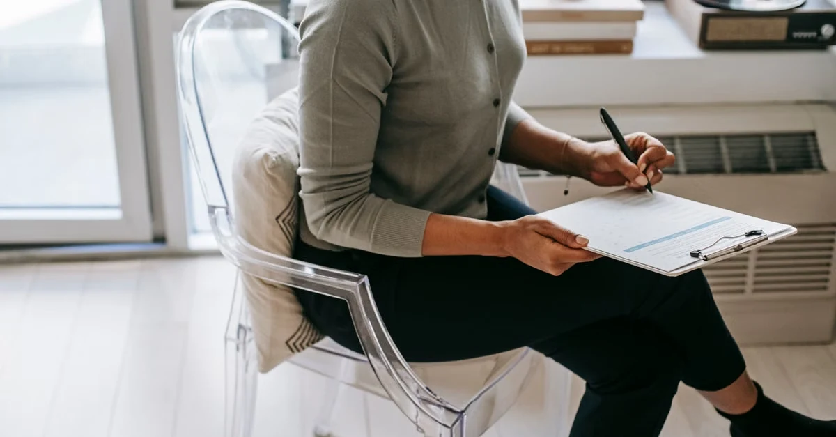 Person in modern office chair writing on clipboard near window, demonstrating executive assistant health coordination.