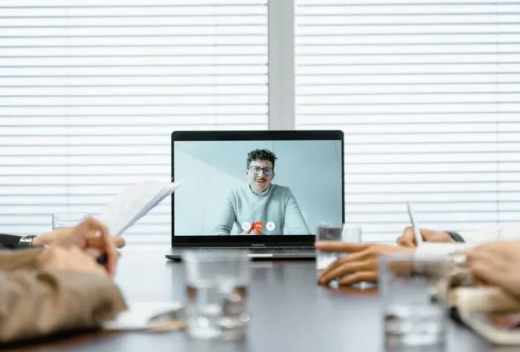 Team members in conference room watching remote executive assistant on laptop screen during virtual meeting.