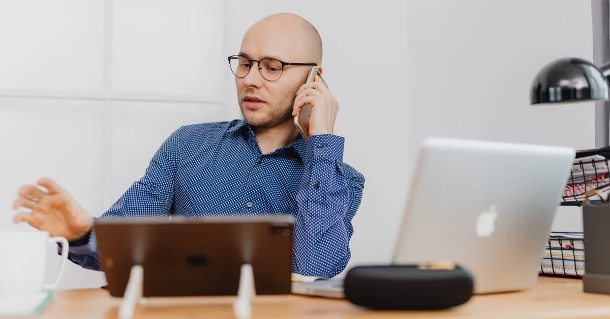 Man in blue shirt on phone call at desk with laptop and tablet, managing remote executive assistant time zones.