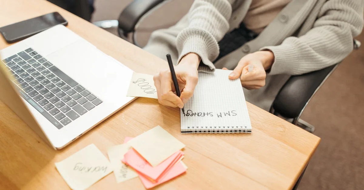 Executive assistant founder writing notes at desk with laptop and sticky notes nearby.