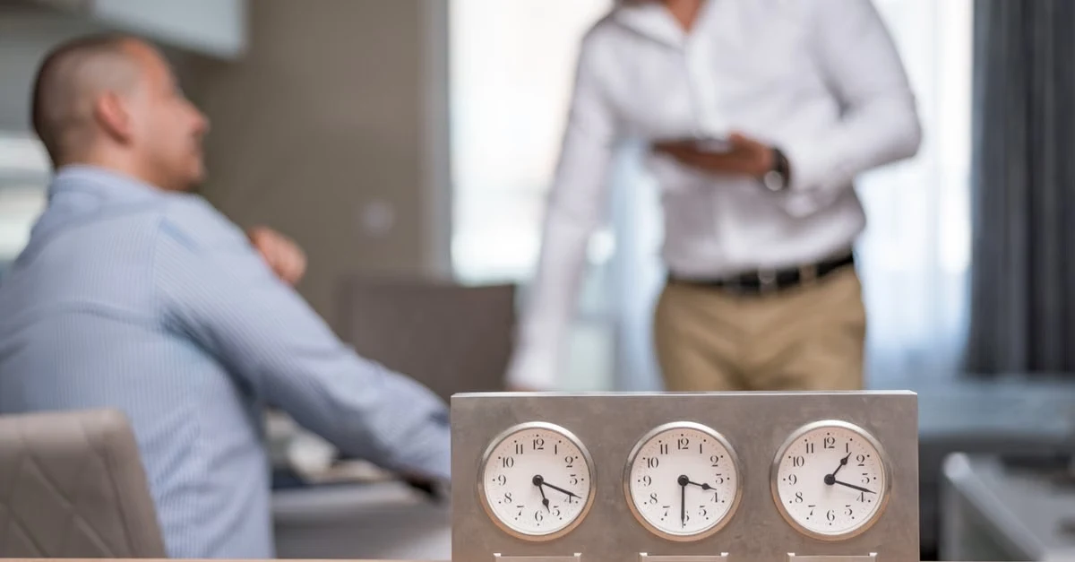 Three clocks showing different times on desk while remote executive assistant and manager discuss time zone scheduling.