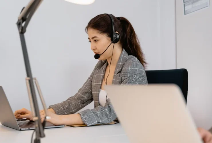 Executive assistant in headset managing decision log on laptop at organized desk with task lamp.