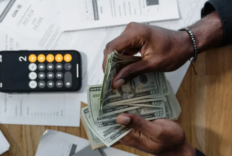 Hand holding cash above calculator and financial documents on desk, representing executive assistant salary costs.