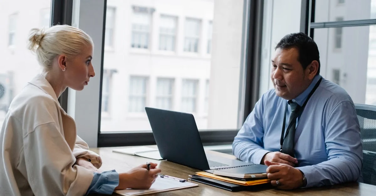 Two professionals discussing hiring strategy at desk with laptop and documents in modern office with city views.