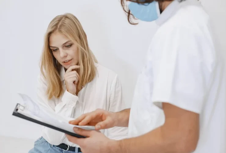 Woman reviewing clipboard with healthcare professional in white coat and mask during consultation.